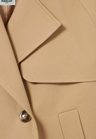 Blazer beige avec un col cranté, des revers structurés, une poche avant et un bouton métallique. Tissu texturé avec des détails de couture visibles.