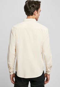 Long-sleeve, cream-colored shirt with a textured finish, buttoned cuffs, and a curved hem, viewed from the back.