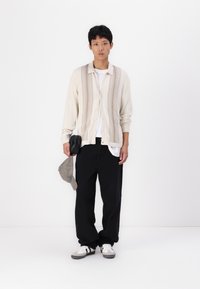 Light beige cardigan with vertical ribbed design, worn over a white shirt, paired with relaxed-fit black pants and gray sneakers. Holding a cap.