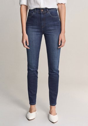 Vrouw die donkere blauwe high-waist skinny jeans draagt, gecombineerd met een wit shirt met lange mouwen en witte instapschoenen, staande op een lichtgrijze vloer.