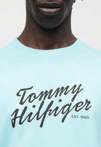 Άνδρας που φοράει ανοιχτή μπλε μπλούζα με μαύρη καλλιγραφική επιγραφή "Tommy Hilfiger" και μικρό κείμενο "EST. 1985" στο στήθος.