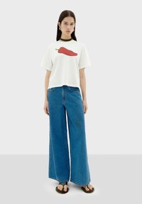 Witte t-shirt met een rode chilipeper graphic, gecombineerd met wijde blauwe denim jeans. Gelaagde sandalen met een harige bovenkant maken de look af.