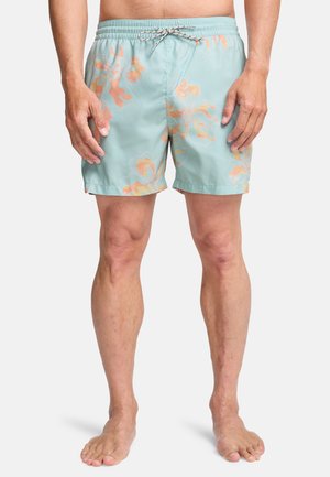 Mann iført lyseblå badeshorts med oransje blomsterprint og snøring i livet, stående barbent mot en ensfarget bakgrunn.