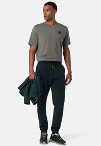 Graues T-Shirt mit kurzen Ärmeln und Logo, kombiniert mit dunkelgrünen Jogginghosen mit Kordelzug. Das Modell hält eine passende Jacke und trägt schwarze Sneakers.