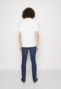 Camiseta blanca de algodón con manga corta combinada con vaqueros ajustados de color azul oscuro y zapatillas negras, vista desde atrás.
