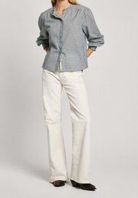 Blusa a cuadros en azul y blanco con mangas abullonadas, cierre de botones y cuello redondo, combinada con jeans blancos acampanados y zapatos oscuros.