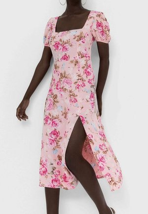 Vrouw draagt een roze midi-jurk met bloemenprint, pofmouwen, een vierkante halslijn en een voorste split, staand tegen een effen witte achtergrond.