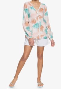 Blusa leggera a maniche lunghe con motivo di foglie di palma rosa e verde acqua, scollatura a V e chiusura con bottoni. Indossata con pantaloni corti bianchi.