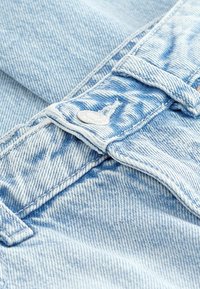 Světle modré džíny z denimu mají stříbrný kovový knoflík, bílé švy a texturovaný povrch s viditelnými jemnými vybledlými vzory.