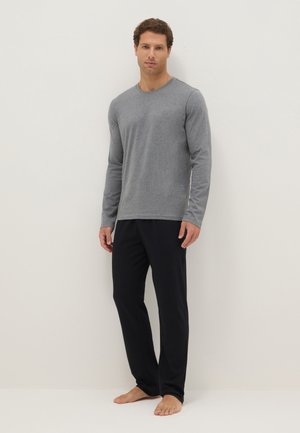 Hombre de pie descalzo con una camiseta gris de manga larga y pantalones negros contra un fondo blanco liso.