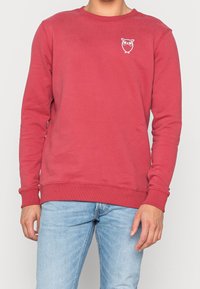 Red cotton sweatshirt with a round neck, ribbed cuffs, and hem. Features a small white owl graphic on the chest. Worn with light blue jeans.