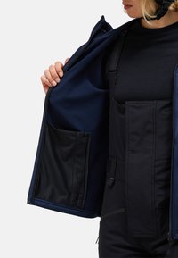 Navy blue jacket with a fleece interior, zippered front, and a side pocket. The garment features adjustable straps and is worn over dark clothing.