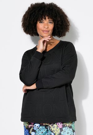 Schwarzer Kapuzenpullover mit strukturiertem Schriftzug, langen Ärmeln und gerippten Bündchen. Kombiniert mit bunten, gemusterten Hosen, die Totenköpfe und Blumen zeigen.