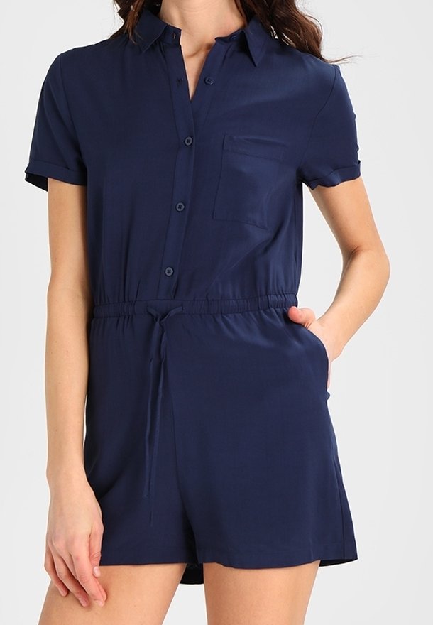 Femme portant une combinaison courte bleu marine à manches courtes boutonnée avec une poche poitrine et une ceinture élastique, main dans la poche latérale.