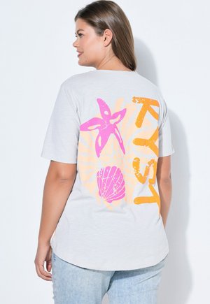 Kobieta w jasnoszarej koszulce z różową rozgwiazdą i grafiką muszli oraz pomarańczowym napisem na plecach, ubrana w jasnoniebieskie dżinsy.