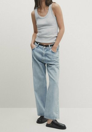 Vrouw die een grijze tanktop draagt, losse lichtblauwe spijkerbroek met zwarte riem, en zwarte instapschoenen, staand met handen in haar zakken.