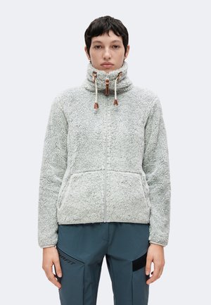 Person trägt eine hellgraue Fleecejacke mit hohem Kragen und Kordelzug, kombiniert mit dunkelblauen Hosen mit schwarzen Aufnähern und Text auf der Tasche.