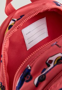 Intérieur d'un sac à dos rouge avec une texture lisse, comportant une étiquette avec un nom et un numéro de téléphone. Des motifs de véhicules colorés sont visibles sur la doublure.
