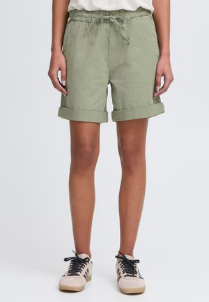 Shorts en lin verts avec une taille élastique et un cordon de serrage, dotés de poignets retroussés et d'une coupe décontractée. Associés à des baskets de couleur claire.