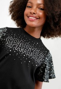 Melna T-krekls ar sudraba krāsā uzrakstītu "SPARKLE", kuru papildina sekvīnu rotājumi uz piedurknēm. Gluda auduma virsma ar brīvu piegriezumu.