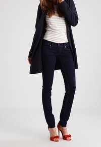 Mörkblå slim-fit jeans, kombinerat med en randig långärmad skjorta och en mörk jacka; röda högklackade sandaler med rosettdetalj.