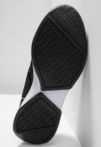 Semelle de sneaker noire avec une surface en caoutchouc texturée, présentant un motif géométrique. Accent blanc près du talon et marqué du logo "TED BAKER".