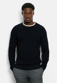 Marineblauer Strickpullover mit strukturiertem Design und geripptem Rundhalsausschnitt, kombiniert mit grauen Hosen. Lange Ärmel und entspannte Passform.