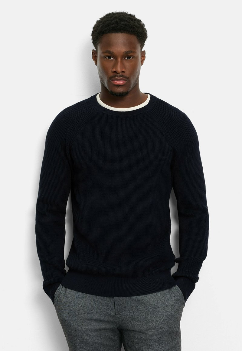 Marineblauer Strickpullover mit strukturiertem Design und geripptem Rundhalsausschnitt, kombiniert mit grauen Hosen. Lange Ärmel und entspannte Passform.