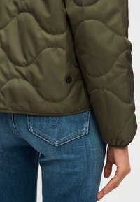 Κοντινό πλάνο προσώπου που φοράει ελιά πράσινο μπουφάν με ραμμένα σχέδια και μπλε τζιν παντελόνι, με εμφανείς λεπτομέρειες χεριού και πίσω τσέπης.