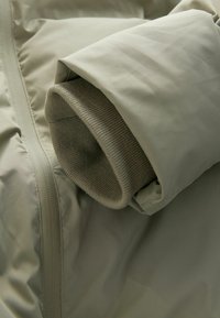 Svetlozelená prešívaná bunda s rukávom a lemom z rebrového pleteného materiálu. Hladký povrch so zipsom a jemným leskom.