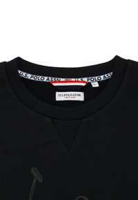 Černé tričko s výstřihem do V. Na přední straně je zelený potisk loga a na límci štítek s nápisem „U.S. POLO ASSN.“