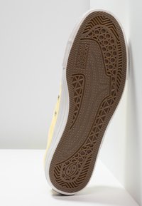 Semelle d'une basket Dockers jaune montrant un motif détaillé de bande de roulement en caoutchouc marron et une semelle intermédiaire blanche sur fond blanc.