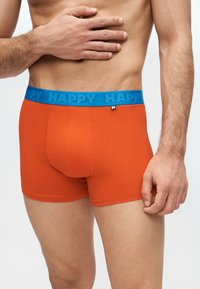 Άνδρας που φοράει φωτεινά πορτοκαλί μποξεράκια με μπλε ζώνη μέσης που αναγράφει "HAPPY", με το χέρι να ακουμπά στο γυμνό στήθος.