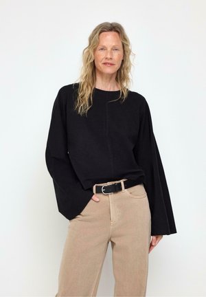 Donna con capelli biondi mossi che indossa un top nero largo a maniche lunghe e pantaloni beige a vita alta con una cintura nera su uno sfondo semplice.