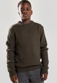 Khakifarbener Ripp-Strickpullover mit langen Ärmeln, Rundhalsausschnitt und strukturiertem Strickdesign, kombiniert mit dunkelgrauen Hosen.
