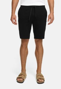 Hombre que lleva pantalones cortos negros con cordón y sandalias beige de doble tira, de pie con los brazos relajados frente a un fondo blanco liso.