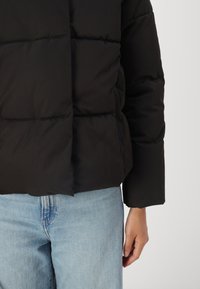 Zwarte pufferjack met een gestikt ontwerp, hoge kraag en lange mouwen, gecombineerd met lichtblauwe denim broeken. Geen zichtbare sluitingen.