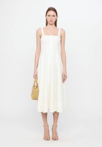 Vestido midi blanco sin mangas con escote cuadrado, cierre frontal de botones y tejido suave. La modelo sostiene un bolso de mimbre, de pie contra un fondo simple.