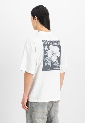 Mann mit langen dunklen Haaren, der ein weißes T-Shirt mit einer schwarz-weißen Hibiskusblume und dem Schriftzug "HAWAII" auf der Rückseite trägt.