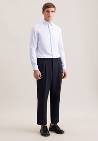 Camicia azzurro chiaro con collo a punta, abbinata a pantaloni blu navy a vita alta e scarpe nere stringate, in piedi su uno sfondo neutro.