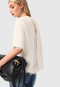 Beige korte mouwtop met kanten rand aan de mouwen en achterkant, gecombineerd met een zwarte leren tas met gouden accenten en ritsdetails.