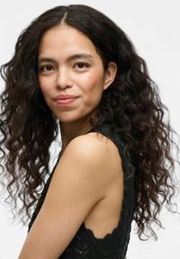 Top de punto negro con un diseño en forma de ondulación, sin mangas, que muestra los hombros y los brazos. El cabello es oscuro, rizado y cae más allá de los hombros.