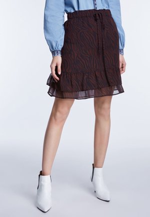 Kobieta ubrana w ciemnoczerwoną i czarną spódnicę w zwierzęcy wzór z falbaną na dole, jasnoniebieską bluzkę z haftowanymi mankietami i białe botki.