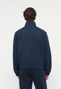 Giacca blu navy con colletto alto, polsini e vita elasticizzati. Texture liscia con un design semplice, senza motivi o hardware visibili.