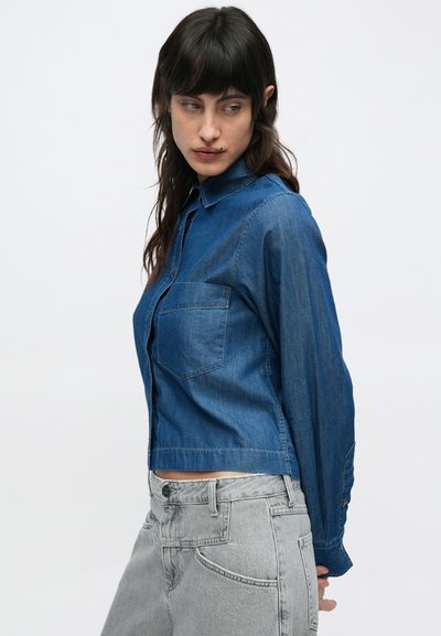 Chemise en jean bleu foncé avec devant boutonné, col et une poche poitrine ; portée avec un jean taille haute gris. Texture lisse.