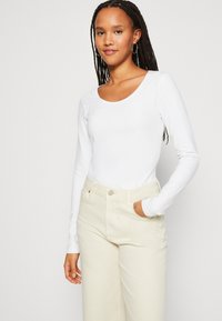 Hvit langermet topp med rund hals, brukt med lys beige høytlivsbukser. Slitt tekstur, standard passform, ingen synlige mønstre.