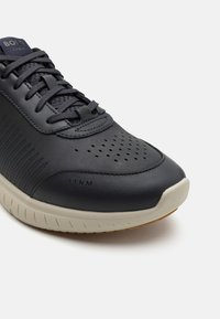 Mørk marineblå skinnsneaker med perforert tåboks, kontrasterende kremgummisåle og teksturert sidepanel. Har flate lisser og logo detalj.