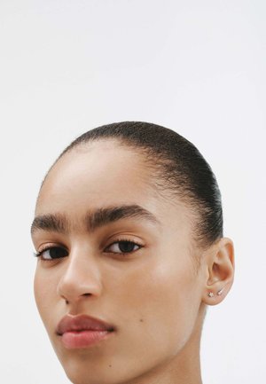 Primer plano de un rostro con piel suave, ojos oscuros y labios naturales. El cabello está peinado hacia atrás. Pequeños pendientes de plata adornan las orejas.