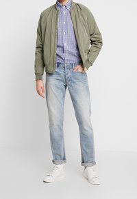 Giacca bomber verde oliva, camicia a scacchi, jeans azzurri e sneakers bianche. Tessuto morbido, vestibilità comoda, risvolti sui jeans.