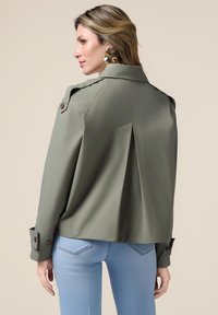 Olijfgroene jas met een getailleerde silhouet, schouderepauletten, knoopmanchetten en een plooidetail aan de achterkant, gecombineerd met blauwe jeans.